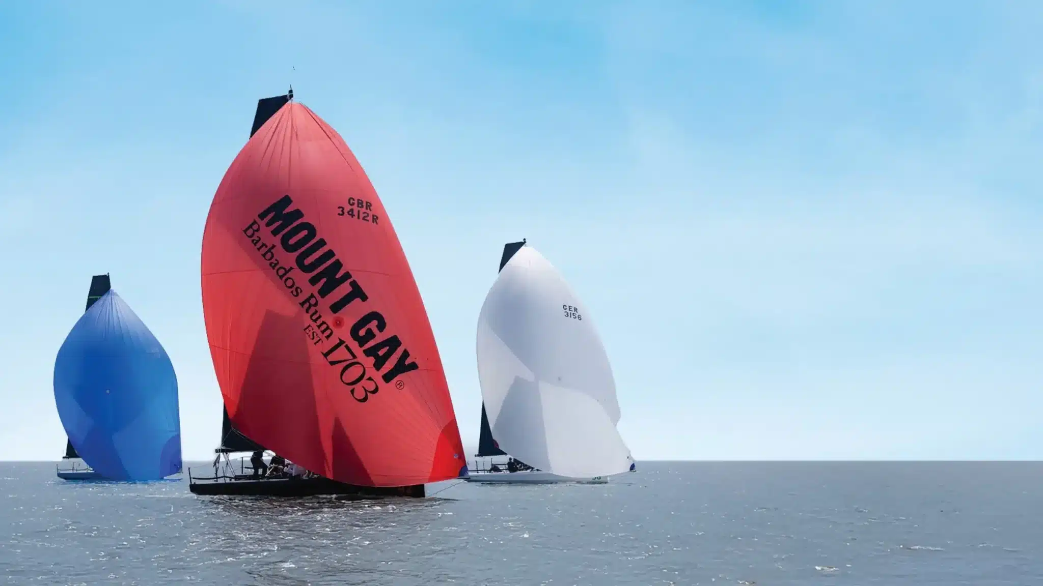 Three racing yachts under spinnaker at Cowes Week, the lead boat flying a red Mount Gay sail.