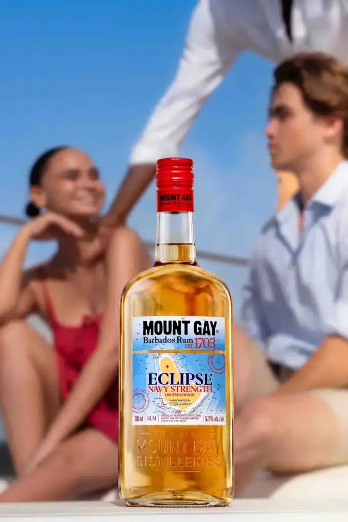 A bottle of Mount Gay Eclipse Navy Strength on deck, with two people relaxing in the sun behind it.