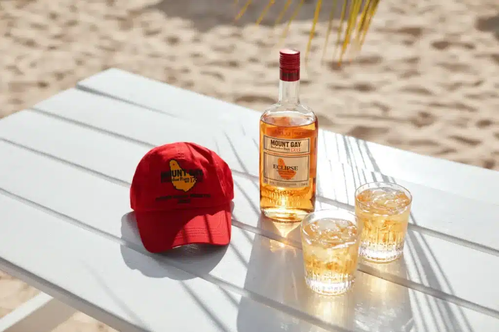 A Cowes Week Regatta Mount Gay Red Cap beside a bottle of Mount Gay Eclipse and two glasses on a beach table.