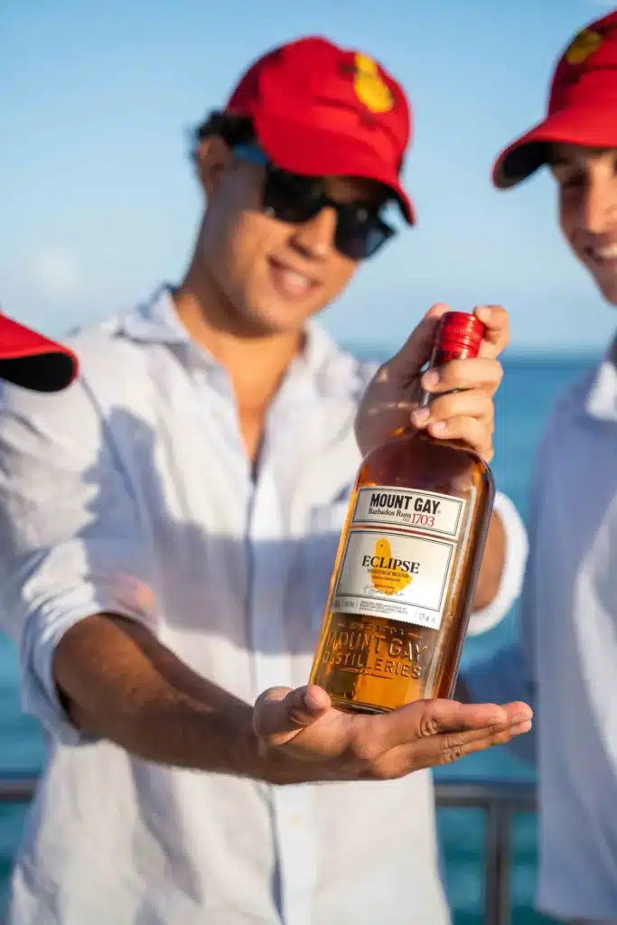 A sailor in a Mount Gay Red Cap holding out a bottle of Mount Gay Eclipse by the sea.